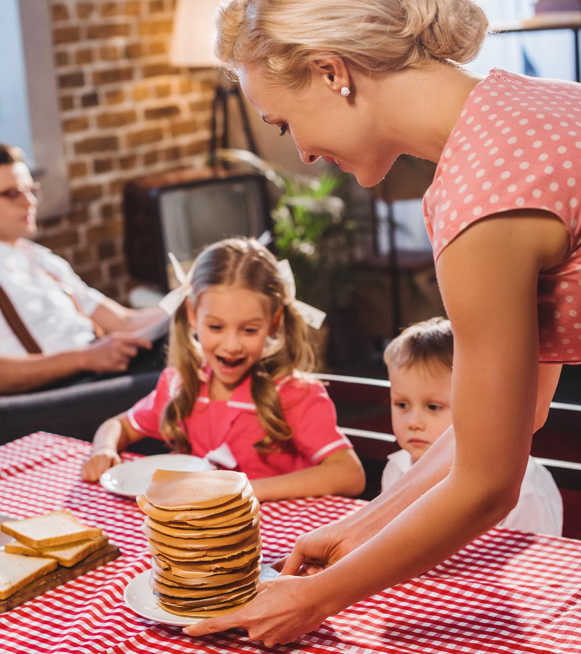 Eine Frau serviert ihren Kindern Pfannkuchen, während der Mann im Hintergrund auf dem Sofa sitzt.