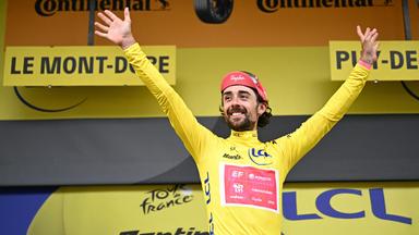 Ben Healy breitet im Gelben Trikot auf dem Podium die Arme aus.
