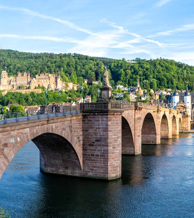 Eine alte Brücke führt über den Neckar. Am anderen Ufer sind eine Schlossruine und die Altstadt von Heidelberg mit einer Kirche zu sehen.