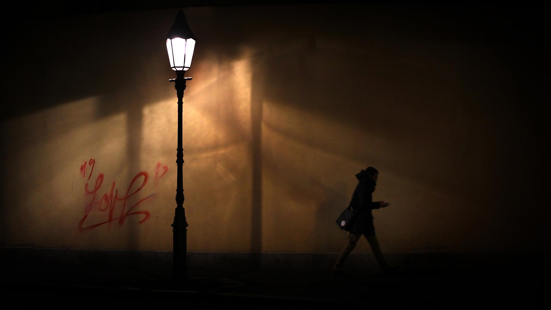 Eine Straßenlaterne erleuchtet einen schmalen Straßenabschnitt in der Nacht. Darunter hastet eine Person auf dem Gehweg vorbei. Sie hält Ihr Smartphone in der Hand.