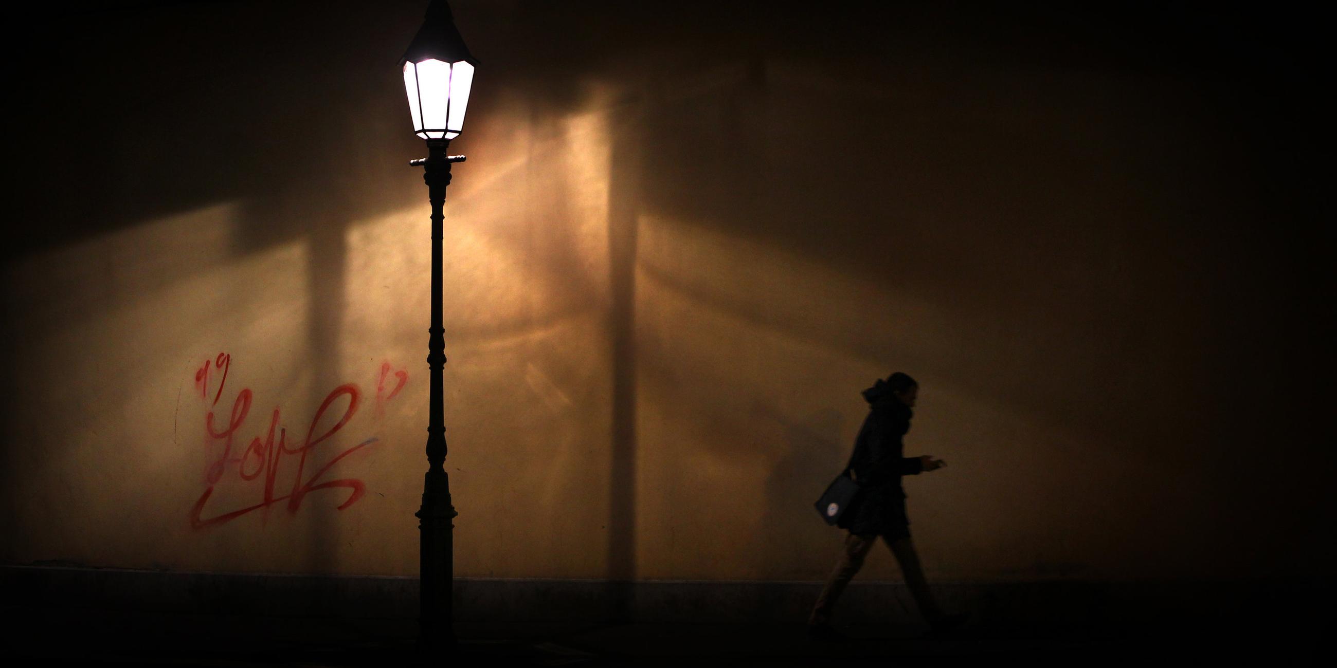 Eine Straßenlaterne erleuchtet einen schmalen Straßenabschnitt in der Nacht. Darunter hastet eine Person auf dem Gehweg vorbei. Sie hält Ihr Smartphone in der Hand.