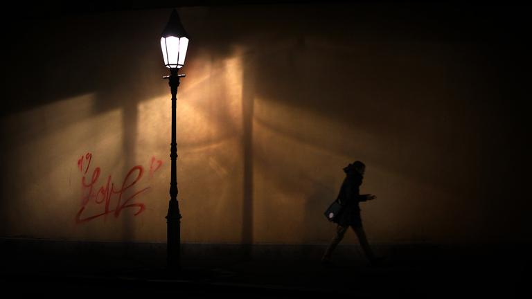 Eine Straßenlaterne erleuchtet einen schmalen Straßenabschnitt in der Nacht. Darunter hastet eine Person auf dem Gehweg vorbei. Sie hält Ihr Smartphone in der Hand.
