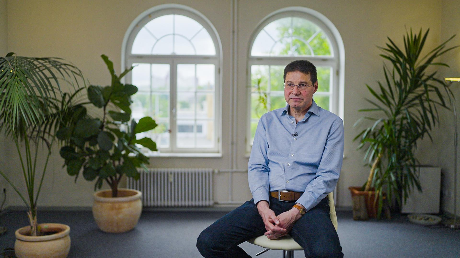 Helge Rettig sitzt in einem blauen Hemd auf einem Hocker. Hinter ihm befinden sich drei große Pflanzen.