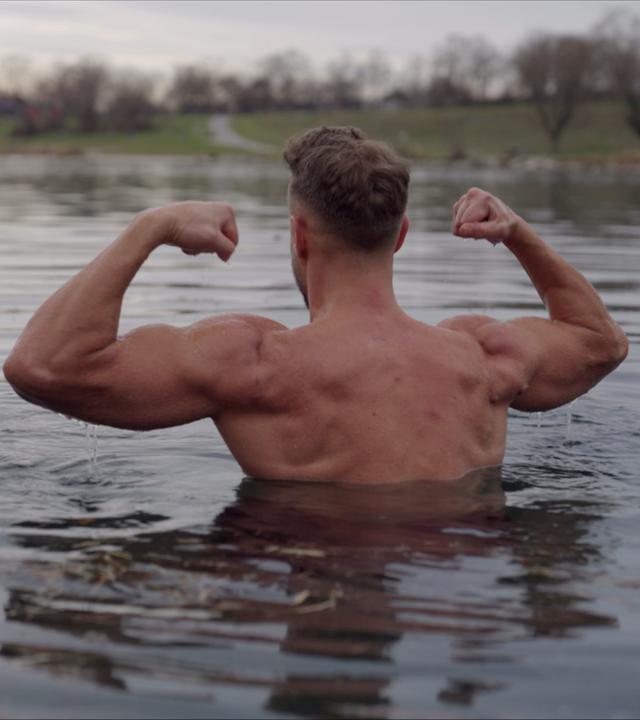 Hendrik beim Eisbaden in der Donau. Er spannt seine Oberarme an und seine Muskeln sind deutlich zu sehen.