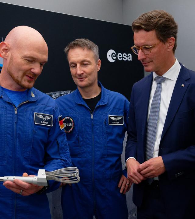 Die Astronauten Matthias Maurer und Alexander Gerst (l) und der nordrhein-westfälische Ministerpräsident Hendrik Wüst (r, CDU) betrachten bei einem Besuch des neuen FLEXhab und der Mondsimulationsanlage LUNA am Deutschen Zentrum für Luft- und Raumfahrt (DLR) und der European Space Agency (ESA) ein Werkzeug zum Aufheben von Mondgestein. 