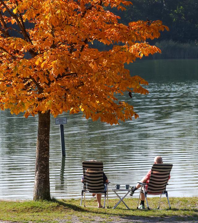 Symbolbild:  Ein Mann und eine Frau sitzen unter einem in herbstlich gefärbten Kastanienbaum an einem See in der Sonne.