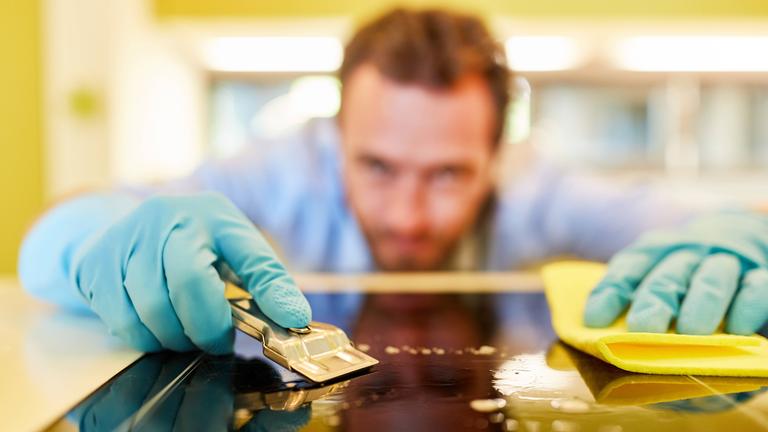 Ein Mann mit Haushaltshandschuhen reinigt ein Glaskeramikfeld mit einer Klinge.