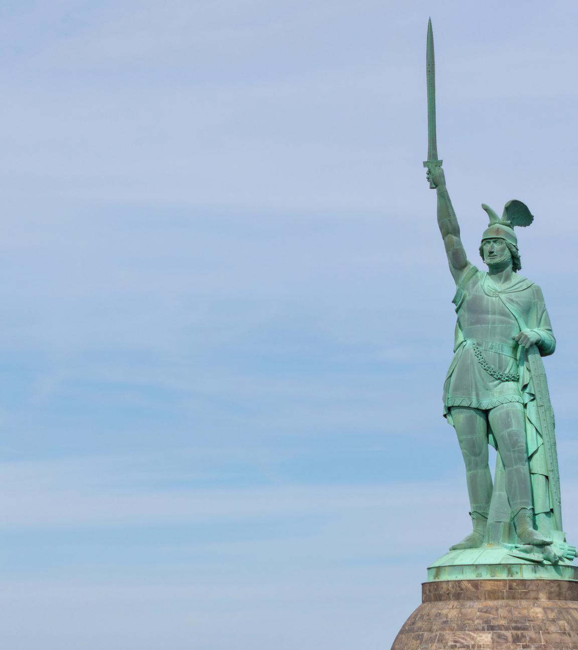 Mit grüner Patina überzogene Statue eines germanischen Kriegers, der einen geflügelten Helm trägt und mit der rechten Hand ein Schwert in den Himmel reckt.