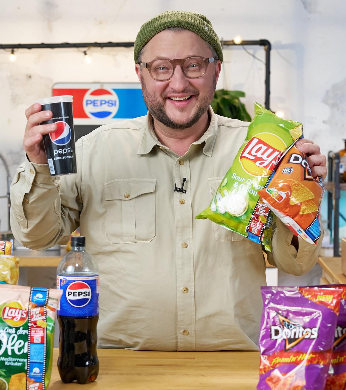 Produktentwickler Sebastian Lege hält einen Becher mit dem Aufdruck "Pepsi" und zwei Chips-Tüten in der Hand, darauf steht "Lays" und "Doritos". Auf dem Tisch vor Sebastian Lege liegen diverse Chipstüten unterschiedlicher Marken, die zum Konzern PepsiCo gehören.