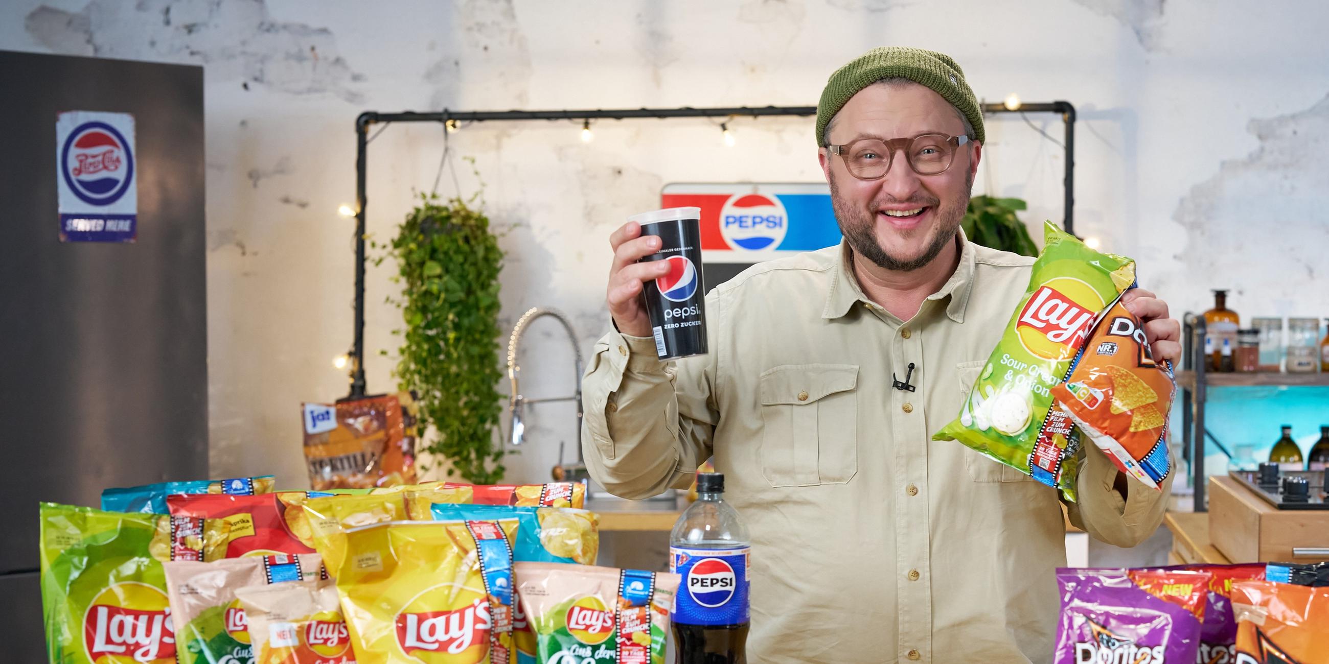 Produktentwickler Sebastian Lege hält einen Becher mit dem Aufdruck "Pepsi" und zwei Chips-Tüten in der Hand, darauf steht "Lays" und "Doritos". Auf dem Tisch vor Sebastian Lege liegen diverse Chipstüten unterschiedlicher Marken, die zum Konzern PepsiCo gehören.