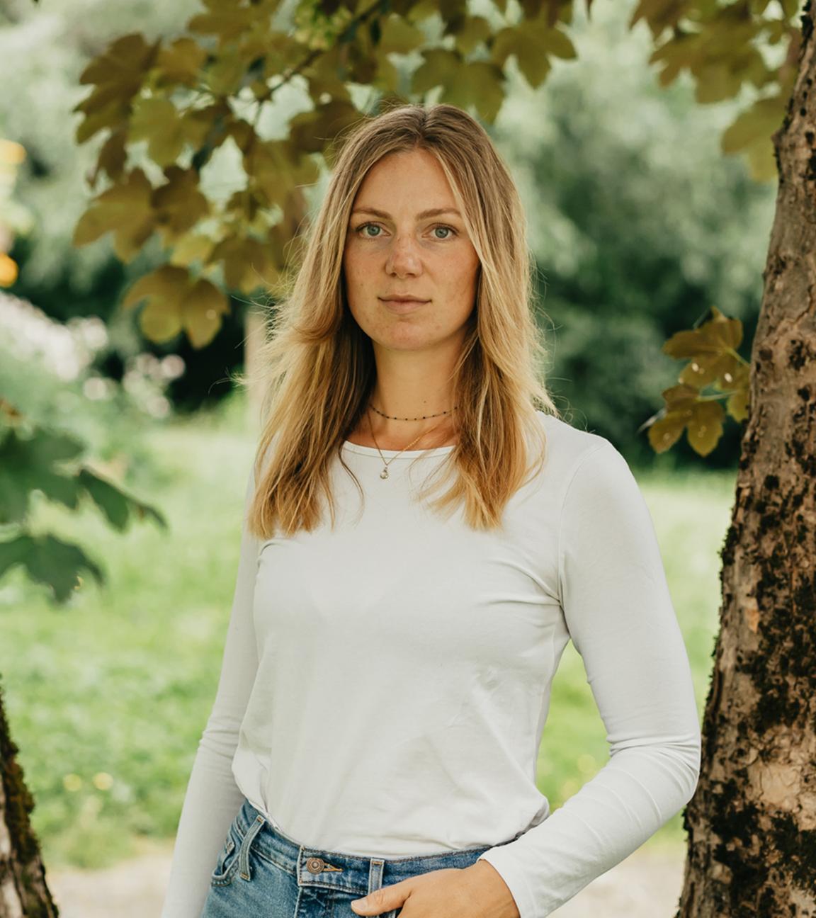 Mara steht in der Natur vor einem Baum und schaut in die Kamera. Eine Hand hat sie leicht in ihre Hosentasche gesteckt. 