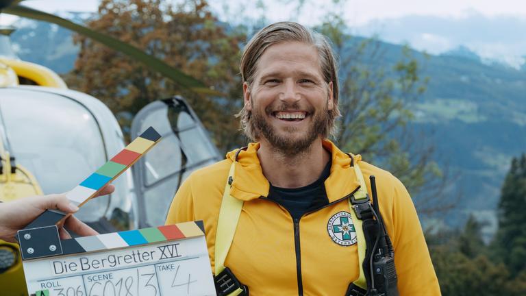 Sebastian Ströbel aus der Serie "Die Bergretter" steht vor einem Rettungs-Helikopter. Vor ihm sieht man eine Filmklappe, denn das Foto entstand während der Dreharbeiten. Die Doku Backstage schaut hinter die Kulissen der Serie.