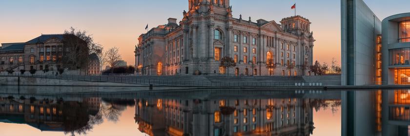 Der Bundestag im Sonnenuntergangslicht. Rechts vom Reichstag ist noch ein Teil des Paul-Löbe-Hauses zu erkennen. Links andere Gebäude im politischen Berlin. Alle Gebäude spiegeln sich am unteren Bildrand in der Spree.