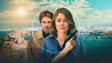 Auftragskillerin Julie Green (Keeley Hawes), die eine Pistole hält, während ihr Sohn Edward Green (Freddie Highmore) dicht hinter ihr steht. Beide blicken mit ernstem Gesichtsausdruck durch das Fadenkreuz eines Zielfernrohrs, das sich über eine sonnige griechische Küstenstadt im Hintergrund legt.