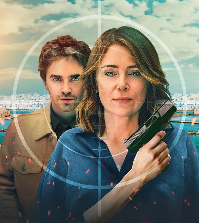 Auftragskillerin Julie Green (Keeley Hawes), die eine Pistole hält, während ihr Sohn Edward Green (Freddie Highmore) dicht hinter ihr steht. Beide blicken mit ernstem Gesichtsausdruck durch das Fadenkreuz eines Zielfernrohrs, das sich über eine sonnige griechische Küstenstadt im Hintergrund legt.