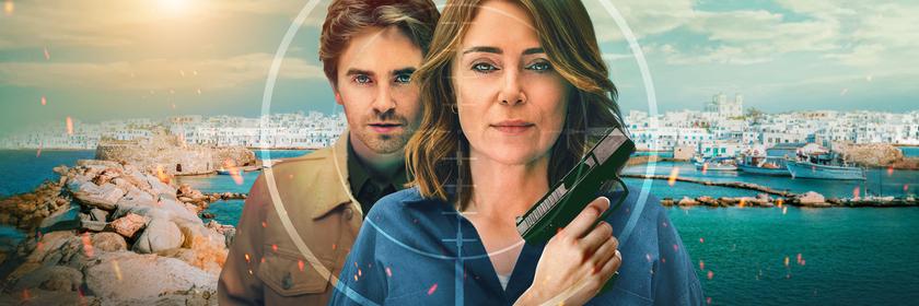 Auftragskillerin Julie Green (Keeley Hawes), die eine Pistole hält, während ihr Sohn Edward Green (Freddie Highmore) dicht hinter ihr steht. Beide blicken mit ernstem Gesichtsausdruck durch das Fadenkreuz eines Zielfernrohrs, das sich über eine sonnige griechische Küstenstadt im Hintergrund legt.
