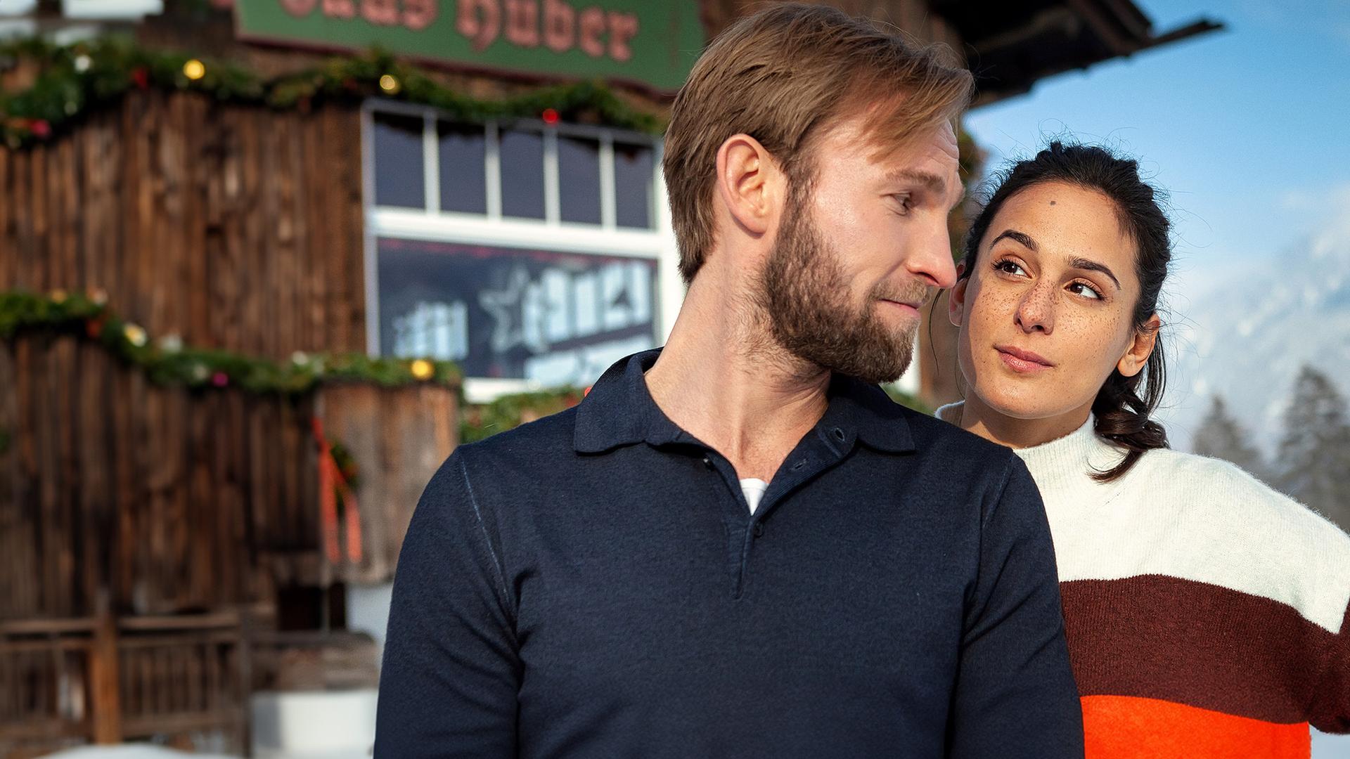Jakob (Jochen Matschke) mit einem blauen Pullover und Alice (Aybi Era) mit einem gestreiften Pullover stehen dicht nebeneinander vor einem Holzhaus. Er blickt sie über die Schulter herausfordernd an, sie hat skeptisch eine Augenbraue hochgezogen. Es liegt Schnee und die Sonne scheint.