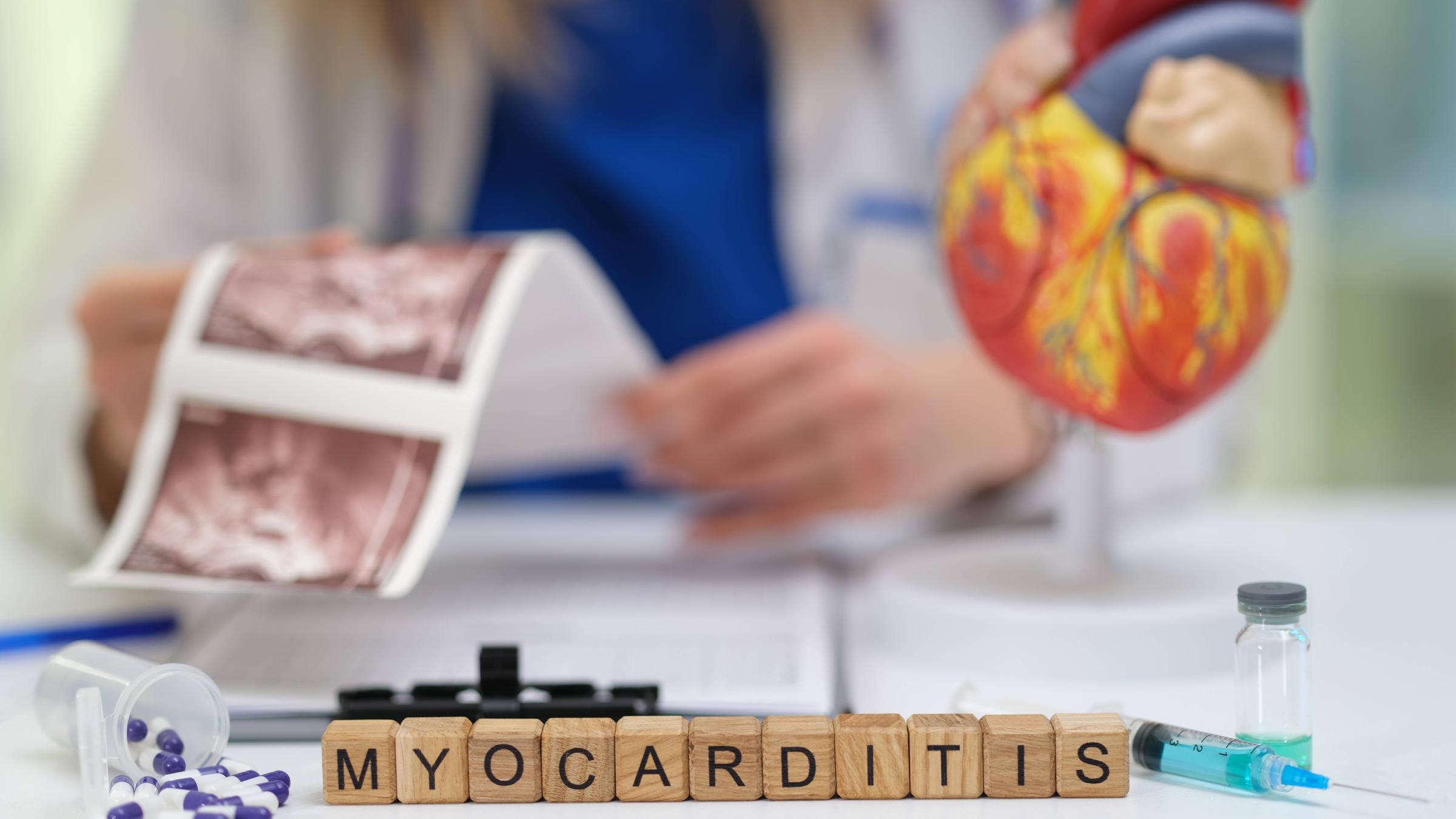 Im Vordergrund ein aus Buchstaben-Steinen gelegter Schriftzug "Myocarditis", dahinter hält einer Person einen Ultraschallbild-Ausdruck vom Herz.