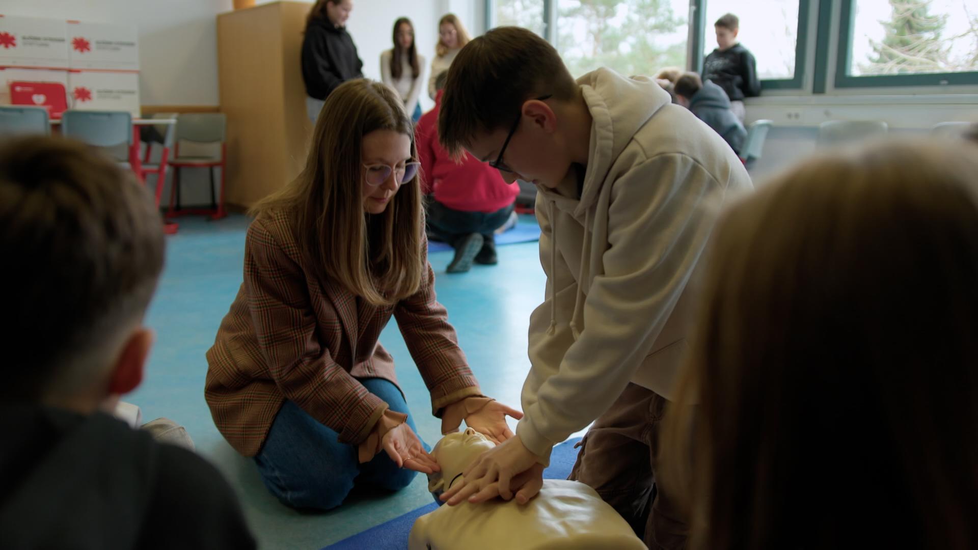 Expertin für "Herzsicherheit an Schulen": Liliane Kozik (links) zeigt einem Schüler (rechts) Reanimationsmaßnahmen an einem Dummy der auf dem Boden liegt.