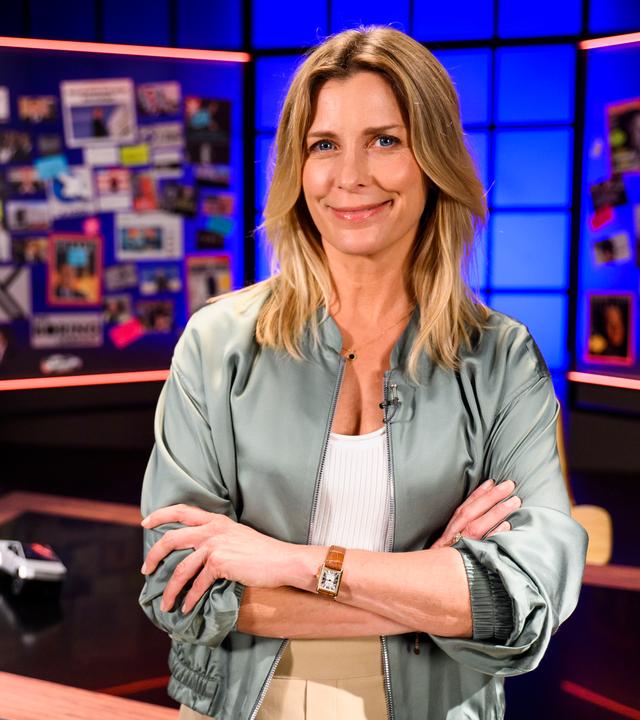 Moderatorin Valerie Niehaus steht mit verschränkten Armen vor einem Schreibtisch. Im Hintergrund befinden sich links und rechts von ihr Glaswände mit Fotografien von Elon Musk und Artikeln über ihn.