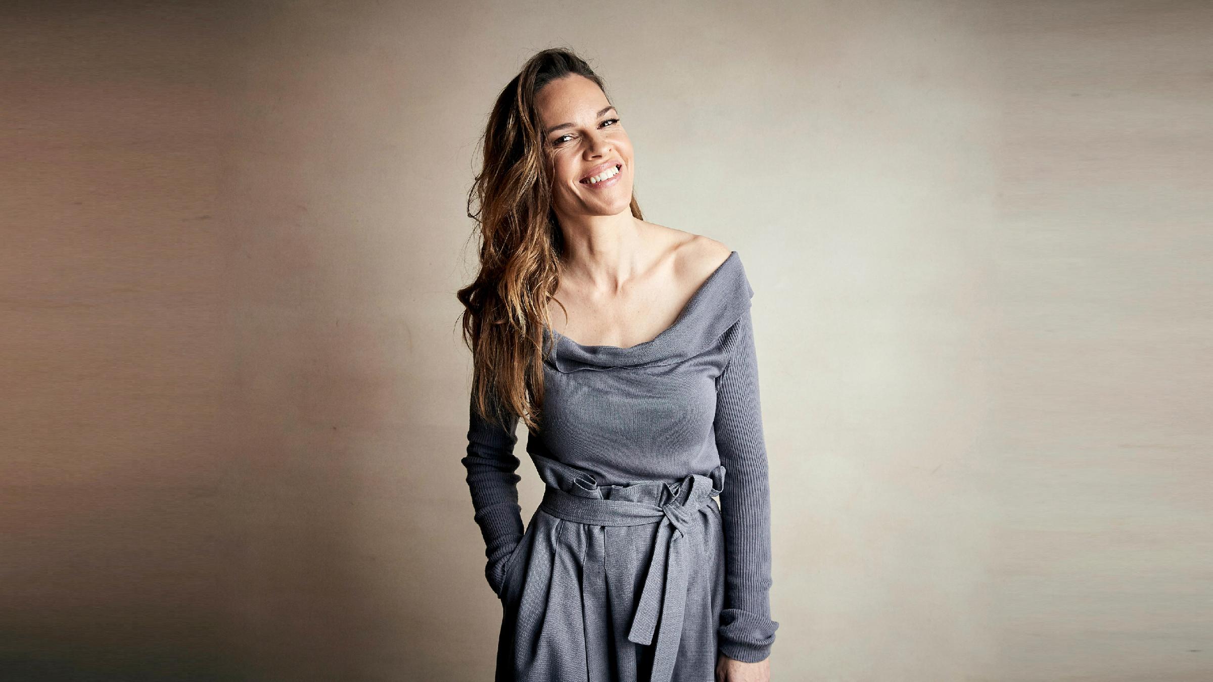 Die US-Schauspielerin Hilary Swank bei einem Fotoshooting. Sie steht vor einem braun-weißen Hintergrund und trägt ein graues Oberteil 