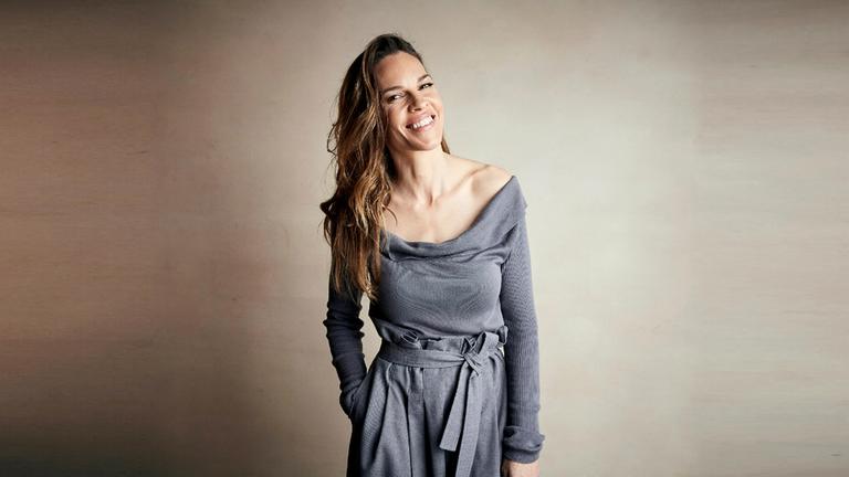Die US-Schauspielerin Hilary Swank bei einem Fotoshooting. Sie steht vor einem braun-weißen Hintergrund und trägt ein graues Oberteil 