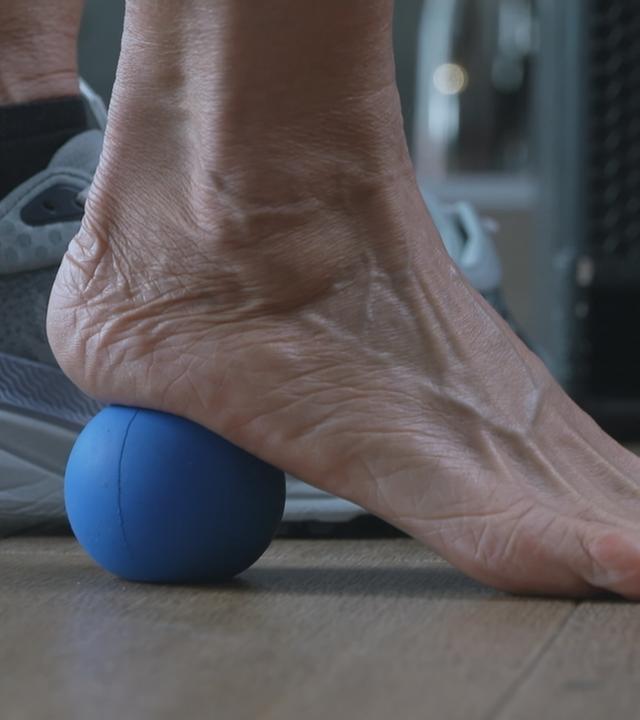 Es sind zwei Füße zu sehen. Der rechte Fuß ist nackt und rollt mit der Ferse auf einem blauen Ball ab. Der linke Fuß ist in einem grauen Schuh.