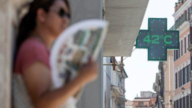 Eine Frau mit Fächer lehnt an einer Hauswand in Rom, auf einer einer Apotheken-Anzeige steht 42 Grad. 