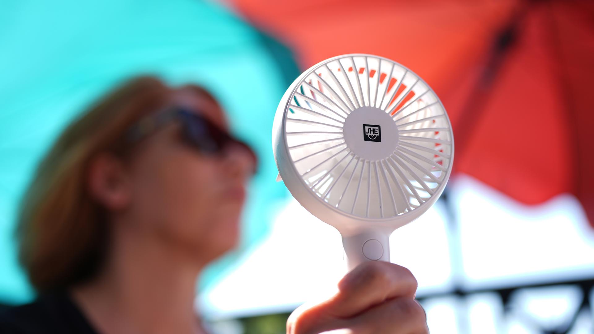 Eine Frau kühlt sich mit einem Handventilator unter Sonnenschirmen.
