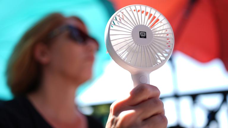 Eine Frau kühlt sich mit einem Handventilator unter Sonnenschirmen.