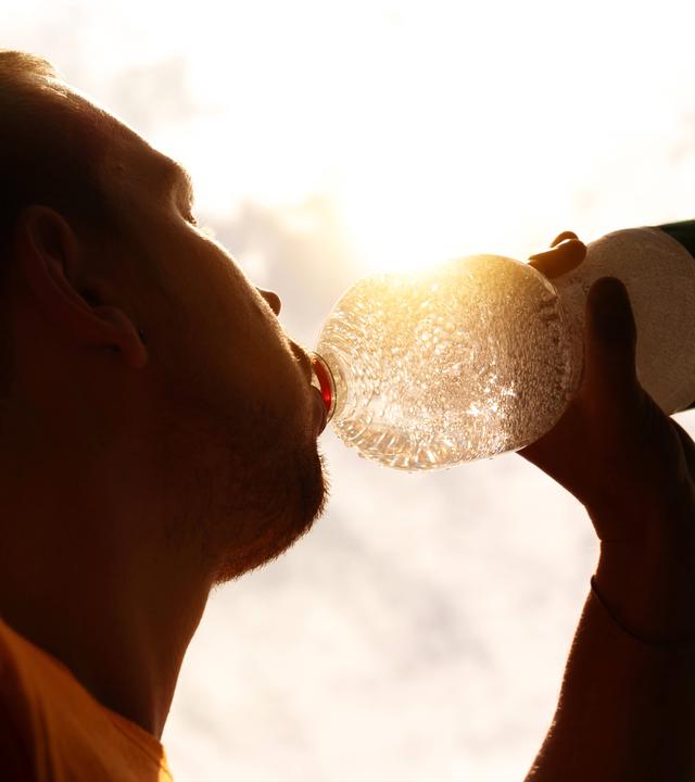 Eine Person trinkt Wasser aus einer transparenten Flasche, die gegen die Sonne gehalten wird.