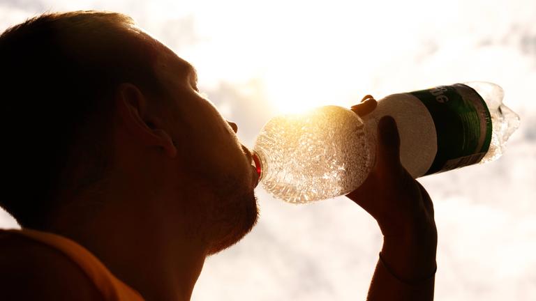 Eine Person trinkt Wasser aus einer transparenten Flasche, die gegen die Sonne gehalten wird.