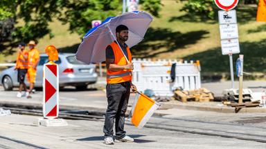 Ein Mitarbeiter steht bei starker Hitze mit einem Sonnenschirm an einer Baustelle.
