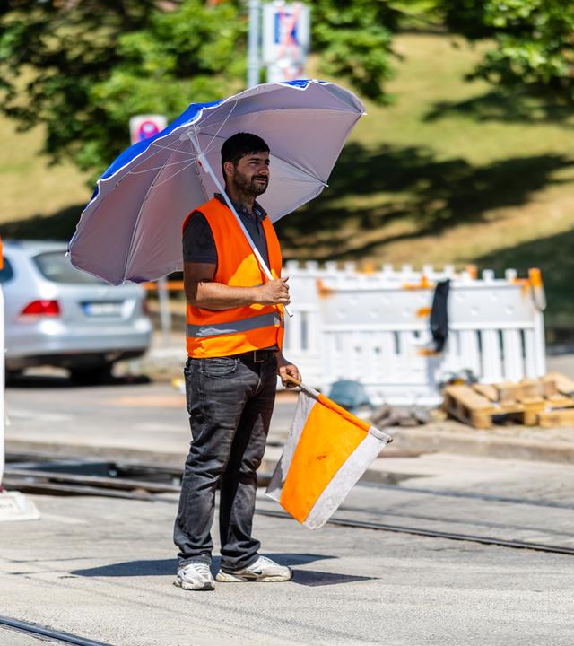Ein Mitarbeiter steht bei starker Hitze mit einem Sonnenschirm an einer Baustelle.