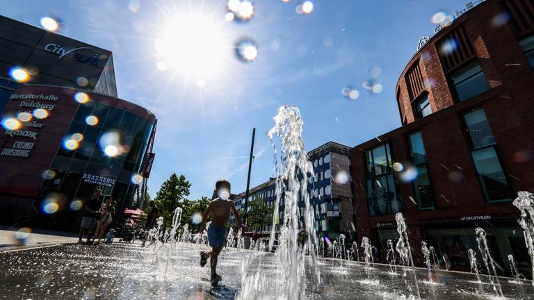 Ein Kind spielt bei bei hoch sommerlichen Temperaturen und strahlendem Sonnenschein in den Wasserstrahlen eines begehbaren Brunnens in der Innenstadt am 26.06.2026.