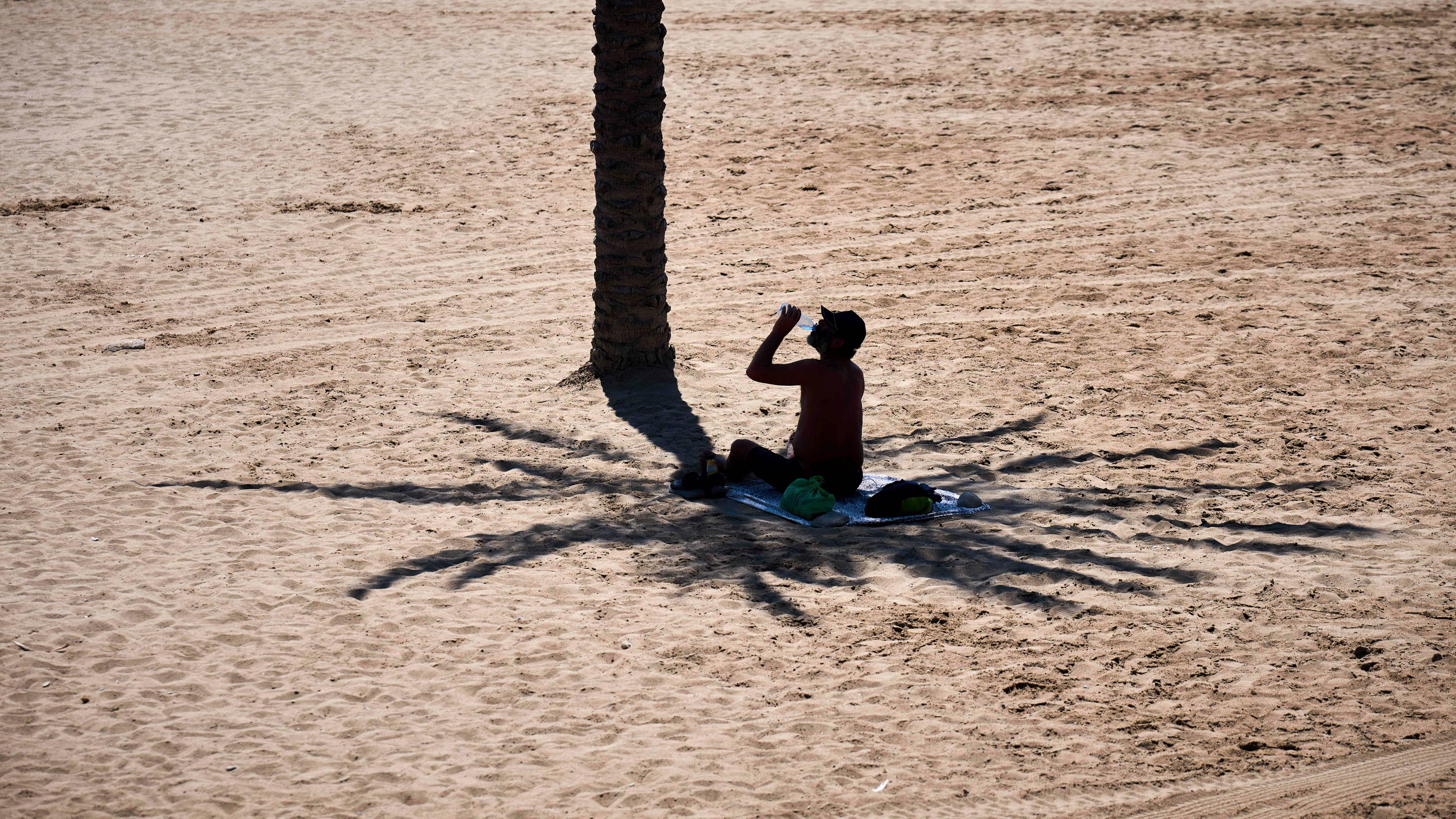 Spanien, Barcelona: Ein Mann nutzt den Schatten einer Palme, um sich vor der Sonne zu schützen, während er an einem heißen Tag am Strand Wasser trinkt.