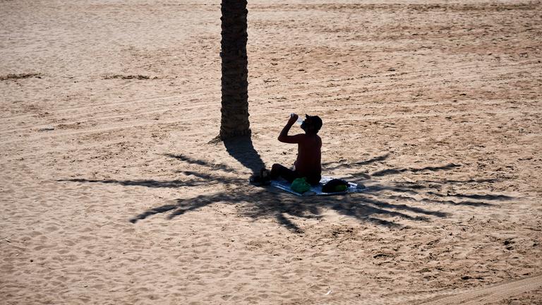 Spanien, Barcelona: Ein Mann nutzt den Schatten einer Palme, um sich vor der Sonne zu schützen, während er an einem heißen Tag am Strand Wasser trinkt.