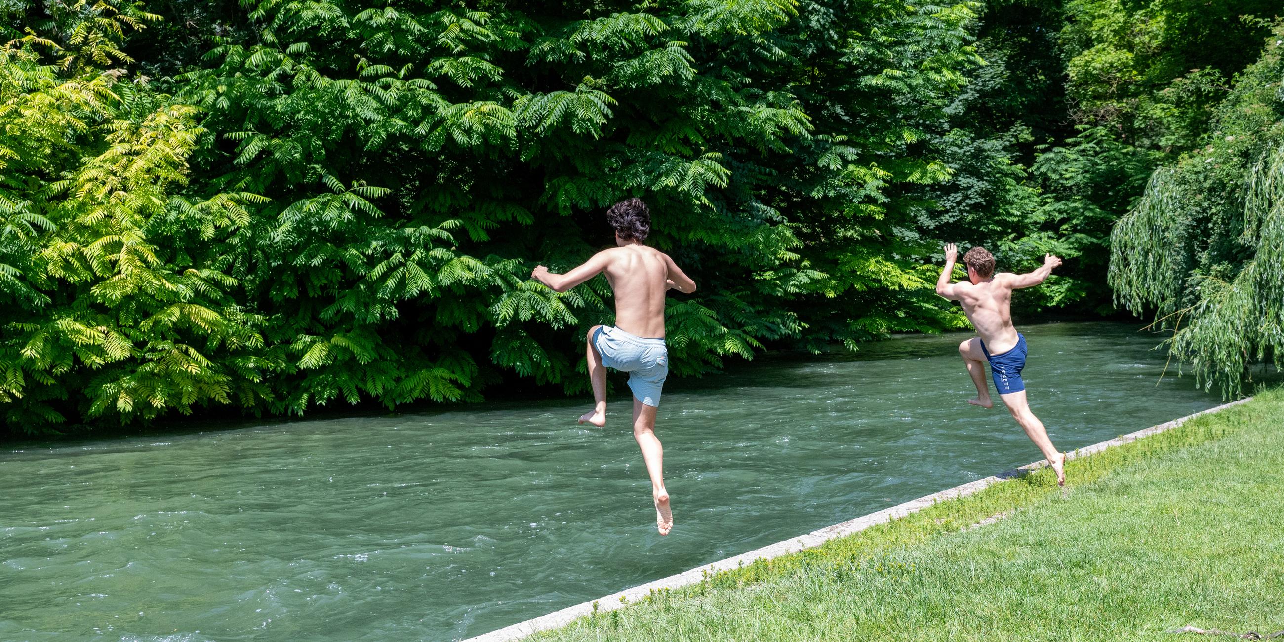 12.06.2025, Bayern, München: Zwei junge Männer springen zur Abkühlung in den Eisbach im Englischen Garten.
