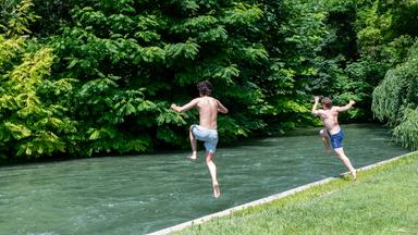 Zwei Kinder springen zur Abkühlung in ein Gewässer im Grünen
