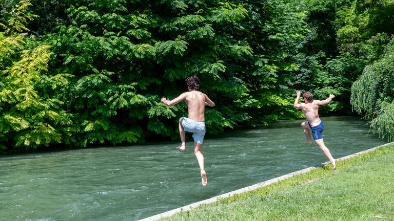 Zwei Kinder springen zur Abkühlung in ein Gewässer im Grünen