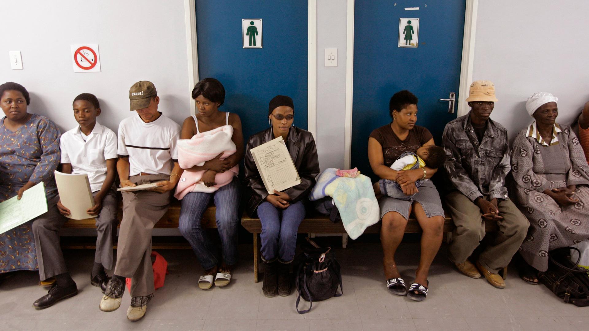 Menschen warten in einem Krankenhaus in Südafrika auf Behandlung. Sie sitzen in einem Flur in einer langen Reihe.