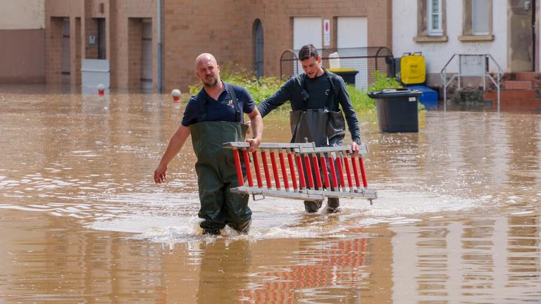 Einsatzkräfte der Freiwilligen Feuerwehr gehen mit einer Leiter durch eine überschwemmte Straße in Kleinblittersdorf im Saarland.
