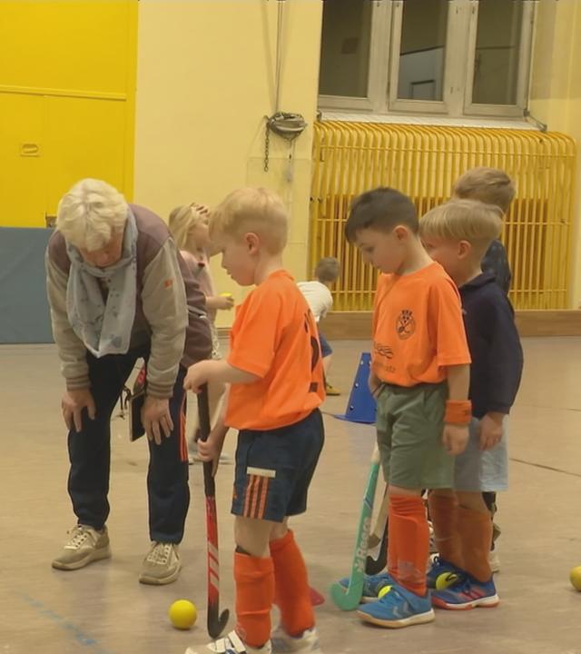Ältere Dame steht mit mehreren Kindern, die Hockey spielen, in einer Sporthalle.