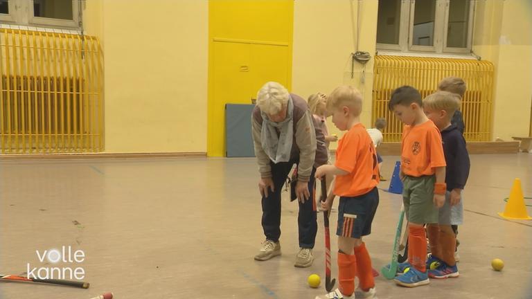 Ältere Dame steht mit mehreren Kindern, die Hockey spielen, in einer Sporthalle.
