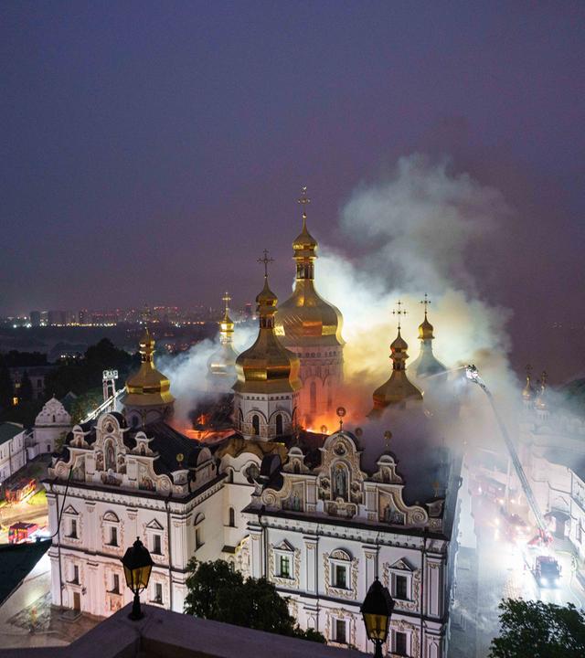 Eine große alte Klosteranlage bestehend aus einem weißen Gebäudekomplex mit goldenen Türmen wird von Feuerschein im Inneren erleuchtet und ist in den Nebel von Löschwasser gehüllt. Rechts und links vom Kloster in den Straßen sind ganz klein Feuerwehrfahrzeuge und Männer mit einer hohen Leiter und Löschschläuchen zu sehen.