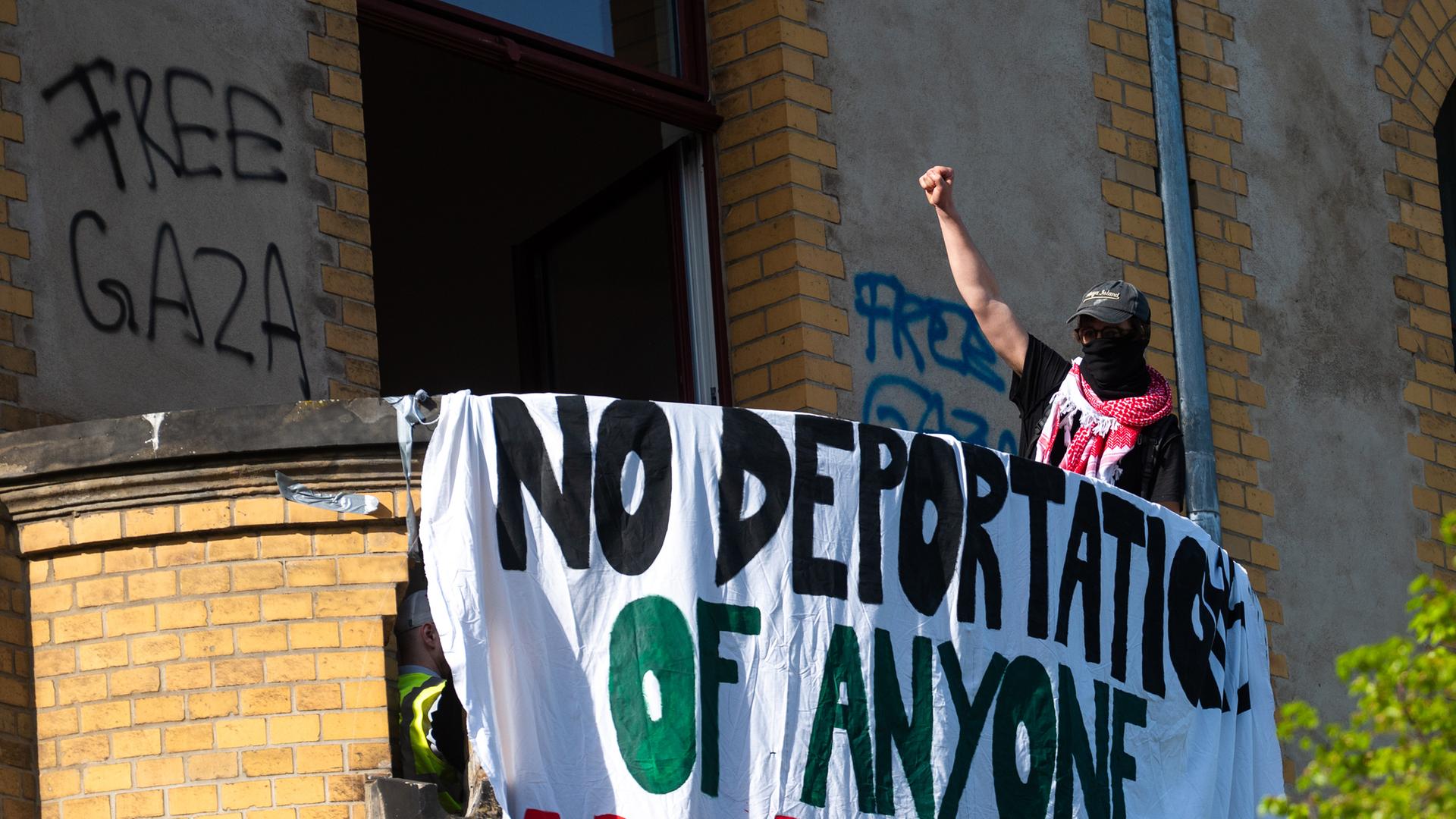 Ein Aktivist hebt die Faust auf einem Balkon in einem Gebäude am Campus Nord der Humboldt-Universität (HU) und steht neben einem Banner mit Forderungen gegen Abschiebungen. 
