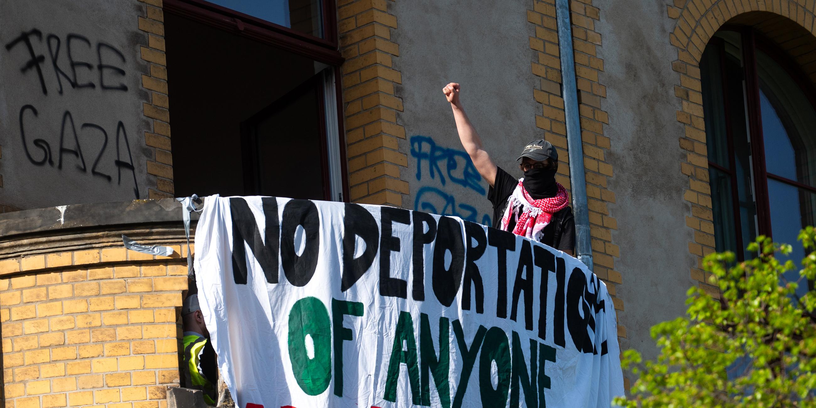 Ein Aktivist hebt die Faust auf einem Balkon in einem Gebäude am Campus Nord der Humboldt-Universität (HU) und steht neben einem Banner mit Forderungen gegen Abschiebungen. 