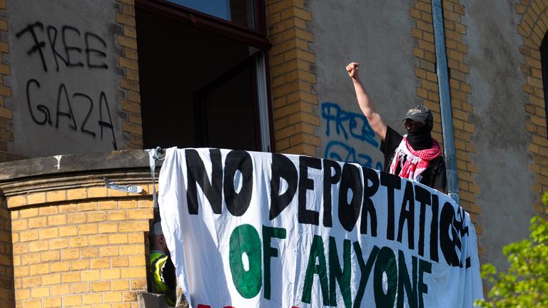 Ein Aktivist hebt die Faust auf einem Balkon in einem Gebäude am Campus Nord der Humboldt-Universität (HU) und steht neben einem Banner mit Forderungen gegen Abschiebungen. 