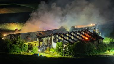 Starker Brand in einem Schweinestall während der Nacht. Über dem Gebäude steigen große Rauchwolken auf, Teile des Dachs sind zerstört.