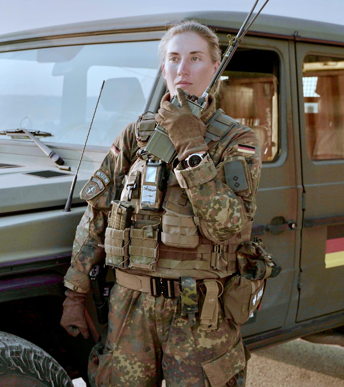 Alina steht in Flecktarn-Uniform und Schutzweste vor einem Bundeswehrauto. Sie hält das Handmikrofon eines Funkgeräts dicht an den Mund und spricht hinein.